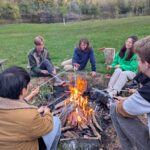 Students sit around a campfire pit roasting marshmallows in a green woody area